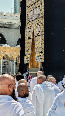 A group of people dressed in white robes gather around the Kaaba, a cube-shaped structure draped in black cloth with gold Arabic script. The scene captures the spiritual atmosphere of a holy place with architectural elements visible in the background.