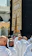 A group of people dressed in white robes gather around the Kaaba, a cube-shaped structure draped in black cloth with gold Arabic script. The scene captures the spiritual atmosphere of a holy place with architectural elements visible in the background.