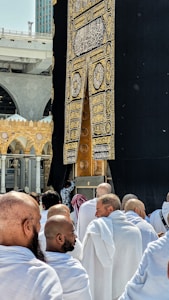 A group of people dressed in white robes gather around the Kaaba, a cube-shaped structure draped in black cloth with gold Arabic script. The scene captures the spiritual atmosphere of a holy place with architectural elements visible in the background.