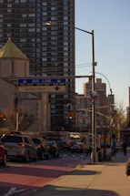 A city street with congested traffic surrounded by tall buildings. The foreground shows several vehicles stopped at a traffic light. A bus lane sign is visible, indicating restricted times for buses. The buildings have a modern design, and a partial view of a dome is visible. Street lights and traffic signs are present, giving the scene an urban feel.