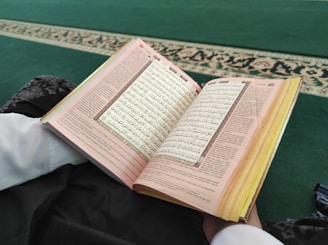 An open book displaying text in a non-Latin script, possibly Arabic, is resting on a fabric surface. A person is partially visible, dressed in white and dark patterned clothing. The background features a green carpet with a decorative border.