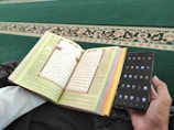 A person is holding a smartphone in one hand and an open book with Arabic text in the other. The pages of the book are colorful at the edges, and the background consists of a green carpet with an ornate design.
