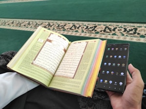 A person is holding a smartphone in one hand and an open book with Arabic text in the other. The pages of the book are colorful at the edges, and the background consists of a green carpet with an ornate design.
