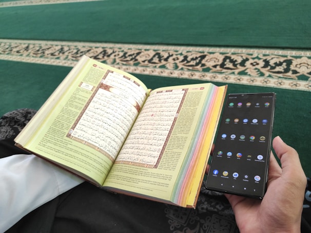 A person is holding a smartphone in one hand and an open book with Arabic text in the other. The pages of the book are colorful at the edges, and the background consists of a green carpet with an ornate design.