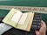A person is holding a smartphone in one hand and an open book with Arabic text in the other. The pages of the book are colorful at the edges, and the background consists of a green carpet with an ornate design.