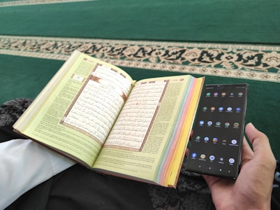 A person is holding a smartphone in one hand and an open book with Arabic text in the other. The pages of the book are colorful at the edges, and the background consists of a green carpet with an ornate design.