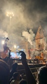 A nighttime religious ceremony with several people in traditional attire performing rituals. Smoke fills the air, and a tall, illuminated temple structure is visible in the background. Several attendees are present, and one person is capturing the scene on a smartphone.