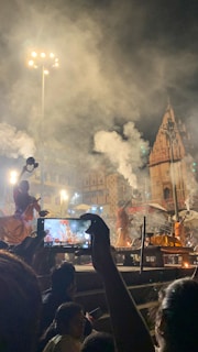 A nighttime religious ceremony with several people in traditional attire performing rituals. Smoke fills the air, and a tall, illuminated temple structure is visible in the background. Several attendees are present, and one person is capturing the scene on a smartphone.