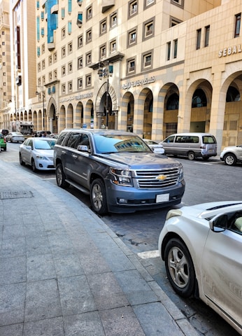 A street scene with several parked cars along a wide sidewalk. The buildings are tall with multiple windows and have Arabic script signage. The street is lined with a mix of modern vehicles, and the architecture has a beige stone facade with arches.