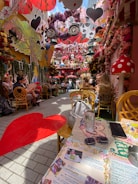 A vibrant outdoor cafe adorned with whimsical decorations inspired by fantasy themes. The area is filled with colorful flowers, oversized mushrooms, and hanging cards. People are seated at tables, enjoying the ambiance. The floor features a red heart design, and a section of the table is visible with a menu, sunglasses, and a smartphone.