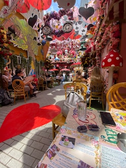 A vibrant outdoor cafe adorned with whimsical decorations inspired by fantasy themes. The area is filled with colorful flowers, oversized mushrooms, and hanging cards. People are seated at tables, enjoying the ambiance. The floor features a red heart design, and a section of the table is visible with a menu, sunglasses, and a smartphone.