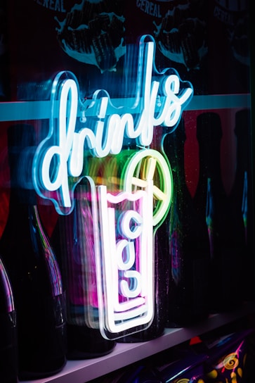 A neon sign with the word 'drinks' in a cursive style is illuminated in blue and white colors. The sign also includes a depiction of a tall cocktail glass with slices of fruit, adding to the vibrant aesthetic. Behind the neon, there are dark bottles creating a contrast and giving depth to the scene.