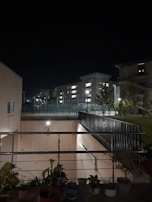 A nighttime outdoor scene featuring residential buildings illuminated by streetlights. There's a fenced area with small trees and a pathway leading to a brightly lit stairwell. Potted plants are visible in the foreground.