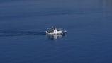 A small white boat with a cabin is moving across calm blue water, leaving a subtle wake. The boat appears to be a fishing vessel, equipped with equipment and antennas.