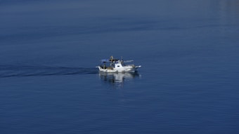 A small white boat with a cabin is moving across calm blue water, leaving a subtle wake. The boat appears to be a fishing vessel, equipped with equipment and antennas.