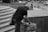 A person with gray hair is bending over, rummaging through a large bag on a city street. The person appears to be wearing a dark t-shirt and jeans. The setting includes stone steps and a busy street with blurred pedestrians and a car. A bicycle is parked in the background.