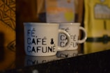 Two white ceramic coffee mugs with black text that reads 'FÉ, CAFÉ & CAFUNÉ' are placed on a shiny black surface. There are blurred coffee packages in the background with distinct patterns and colors, including yellow and brown.