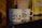 Two white ceramic coffee mugs with black text that reads 'FÉ, CAFÉ & CAFUNÉ' are placed on a shiny black surface. There are blurred coffee packages in the background with distinct patterns and colors, including yellow and brown.