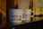 Two white ceramic coffee mugs with black text that reads 'FÉ, CAFÉ & CAFUNÉ' are placed on a shiny black surface. There are blurred coffee packages in the background with distinct patterns and colors, including yellow and brown.