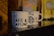 Two white ceramic coffee mugs with black text that reads 'FÉ, CAFÉ & CAFUNÉ' are placed on a shiny black surface. There are blurred coffee packages in the background with distinct patterns and colors, including yellow and brown.