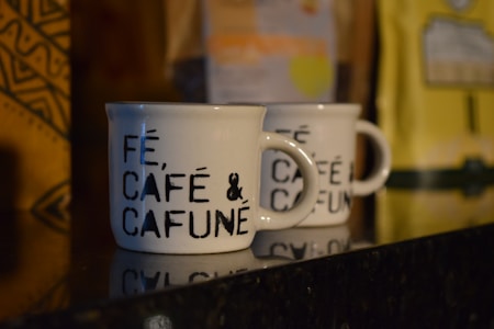 Two white ceramic coffee mugs with black text that reads 'FÉ, CAFÉ & CAFUNÉ' are placed on a shiny black surface. There are blurred coffee packages in the background with distinct patterns and colors, including yellow and brown.