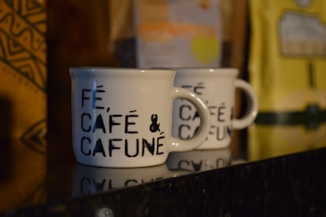 Two white ceramic coffee mugs with black text that reads 'F&Eacute;, CAF&Eacute; & CAFUN&Eacute;' are placed on a shiny black surface. There are blurred coffee packages in the background with distinct patterns and colors, including yellow and brown.