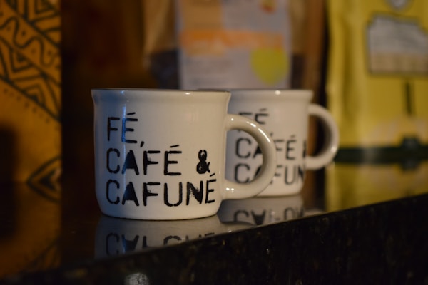 Two white ceramic coffee mugs with black text that reads 'F&Eacute;, CAF&Eacute; & CAFUN&Eacute;' are placed on a shiny black surface. There are blurred coffee packages in the background with distinct patterns and colors, including yellow and brown.