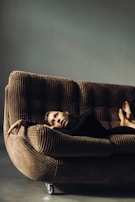 A person is lounging on a corduroy couch with wheels, looking contemplative. The setting is minimalist with muted lighting, creating a soft and introspective atmosphere.