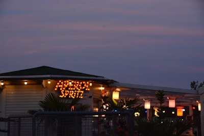 A cozy setting with a sign that reads 'APEROL SPRITZ' in illuminated letters. The area features a canopy adorned with various colored lanterns emitting warm light. Palm plants are visible, adding a tropical ambiance against the backdrop of a dusky sky.