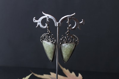 A pair of ornate earrings hang on a decorative metal stand. The earrings feature an intricate swirl design with a large, teardrop-shaped green stone set in a silver-colored metal. The background is dark, emphasizing the refined details of the earrings.