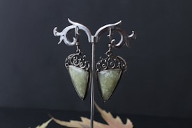A pair of ornate earrings hang on a decorative metal stand. The earrings feature an intricate swirl design with a large, teardrop-shaped green stone set in a silver-colored metal. The background is dark, emphasizing the refined details of the earrings.