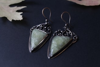 A pair of artisanal earrings featuring geometric green stones set in ornate metal settings. The metal is dark and intricately designed, giving a vintage or bohemian feel. The earrings rest on a dark surface with a muted leaf in the background, enhancing the elegant aesthetic.