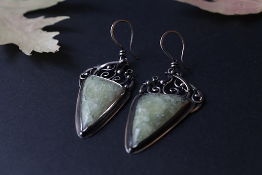 A pair of artisanal earrings featuring geometric green stones set in ornate metal settings. The metal is dark and intricately designed, giving a vintage or bohemian feel. The earrings rest on a dark surface with a muted leaf in the background, enhancing the elegant aesthetic.