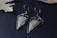 A pair of artisanal earrings featuring geometric green stones set in ornate metal settings. The metal is dark and intricately designed, giving a vintage or bohemian feel. The earrings rest on a dark surface with a muted leaf in the background, enhancing the elegant aesthetic.