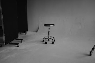 A monochrome photograph of a simple, empty studio space with a single, adjustable stool on casters positioned centrally. The stool has a metallic base with a round, cushioned seat. Large, dark panels on wheels are situated to the left side, with one partially out of frame. The floor and background wall are bare, adding to the minimalistic environment. The lighting is soft, casting slight shadows on the ground.