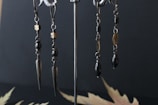 A pair of metal earrings with intricate designs hanging from hooks. The earrings feature dark beads, gold-colored accents, and elongated metallic spikes as pendants. They are displayed against a dark, matte background with blurred leaf-like patterns on the surface.