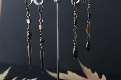 A pair of metal earrings with intricate designs hanging from hooks. The earrings feature dark beads, gold-colored accents, and elongated metallic spikes as pendants. They are displayed against a dark, matte background with blurred leaf-like patterns on the surface.