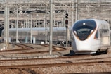 A modern high-speed train navigating a set of tracks in a busy rail yard. The background is filled with numerous metal beams, electrical wires, and railway infrastructure. The train has a sleek, aerodynamic design with a white and silver exterior and orange accents.