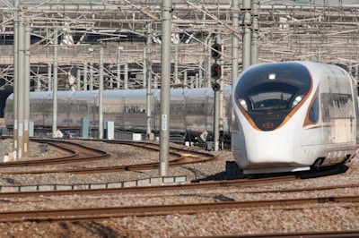 A modern high-speed train navigating a set of tracks in a busy rail yard. The background is filled with numerous metal beams, electrical wires, and railway infrastructure. The train has a sleek, aerodynamic design with a white and silver exterior and orange accents.
