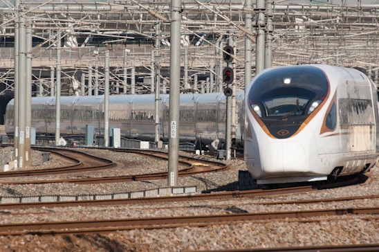 A modern high-speed train navigating a set of tracks in a busy rail yard. The background is filled with numerous metal beams, electrical wires, and railway infrastructure. The train has a sleek, aerodynamic design with a white and silver exterior and orange accents.