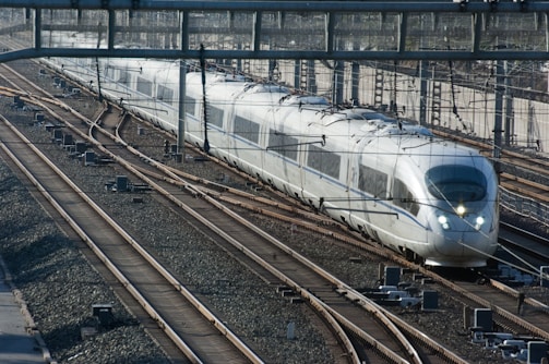 A sleek, modern high-speed train travels along parallel tracks. The train is silver with a streamlined design and large windows. Overhead wires run above the train, which appears to be in motion on a clear day. The setting includes multiple tracks and signaling equipment.