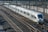 A sleek, modern high-speed train travels along parallel tracks. The train is silver with a streamlined design and large windows. Overhead wires run above the train, which appears to be in motion on a clear day. The setting includes multiple tracks and signaling equipment.