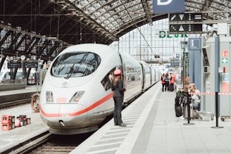 A sleek, modern train is stationed at a railway platform under an expansive, arched glass roof. Several people are visible, including a person in uniform, appearing to interact with the train. In the background, passengers await on the platform. Signage and amenities, such as a clock and information panels, are positioned throughout the station area.