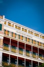 A multi-story hotel building with decorative yellow wrought iron railings and green shutters on the windows. The façade features a combination of light beige, green, and earthy red colors with the name 'Grand Hotel Imperial' prominently displayed. The sky above is clear and deep blue.