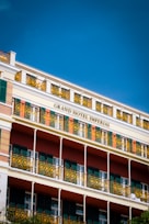 A multi-story hotel building with decorative yellow wrought iron railings and green shutters on the windows. The façade features a combination of light beige, green, and earthy red colors with the name 'Grand Hotel Imperial' prominently displayed. The sky above is clear and deep blue.