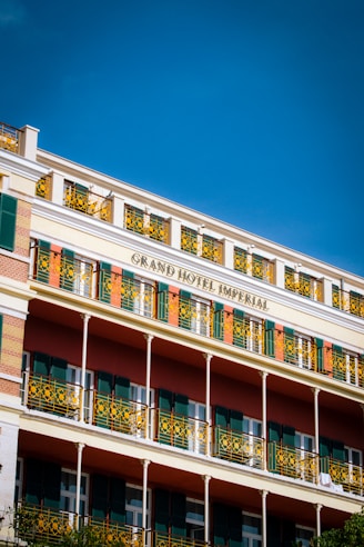 A multi-story hotel building with decorative yellow wrought iron railings and green shutters on the windows. The façade features a combination of light beige, green, and earthy red colors with the name 'Grand Hotel Imperial' prominently displayed. The sky above is clear and deep blue.
