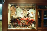 A vintage store display features a bright red motorcycle with a leather saddlebag, surrounded by various apparel items such as hats and patterned shirts hanging in the background. Accessories like gloves and bags are also present, creating an old-fashioned and rustic ambience.