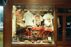 A vintage store display features a bright red motorcycle with a leather saddlebag, surrounded by various apparel items such as hats and patterned shirts hanging in the background. Accessories like gloves and bags are also present, creating an old-fashioned and rustic ambience.