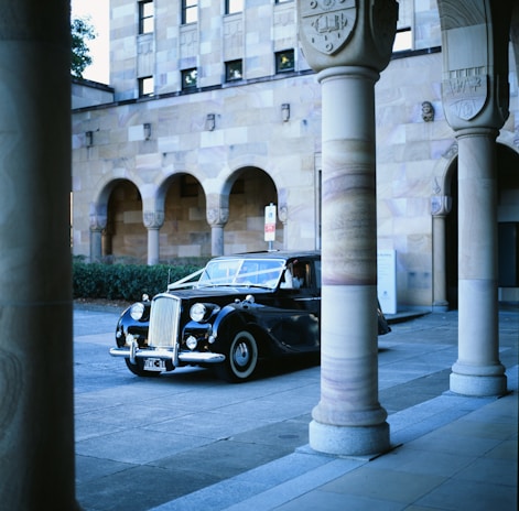 A classic black car is parked in front of an ornate building with textured stone walls and arched windows. The building features intricate columns and decorative elements.