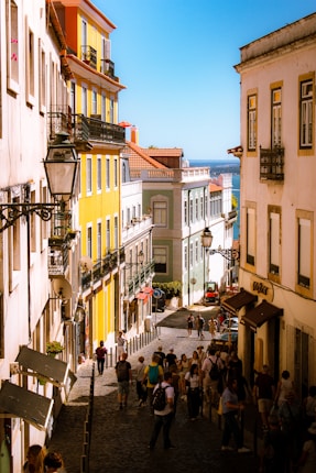 A vibrant street scene with colorful buildings lining a cobblestone road. The architecture features bright yellow and pastel hues. People stroll along the street, enjoying the sunny day, while street lamps and balconies add charm to the setting.